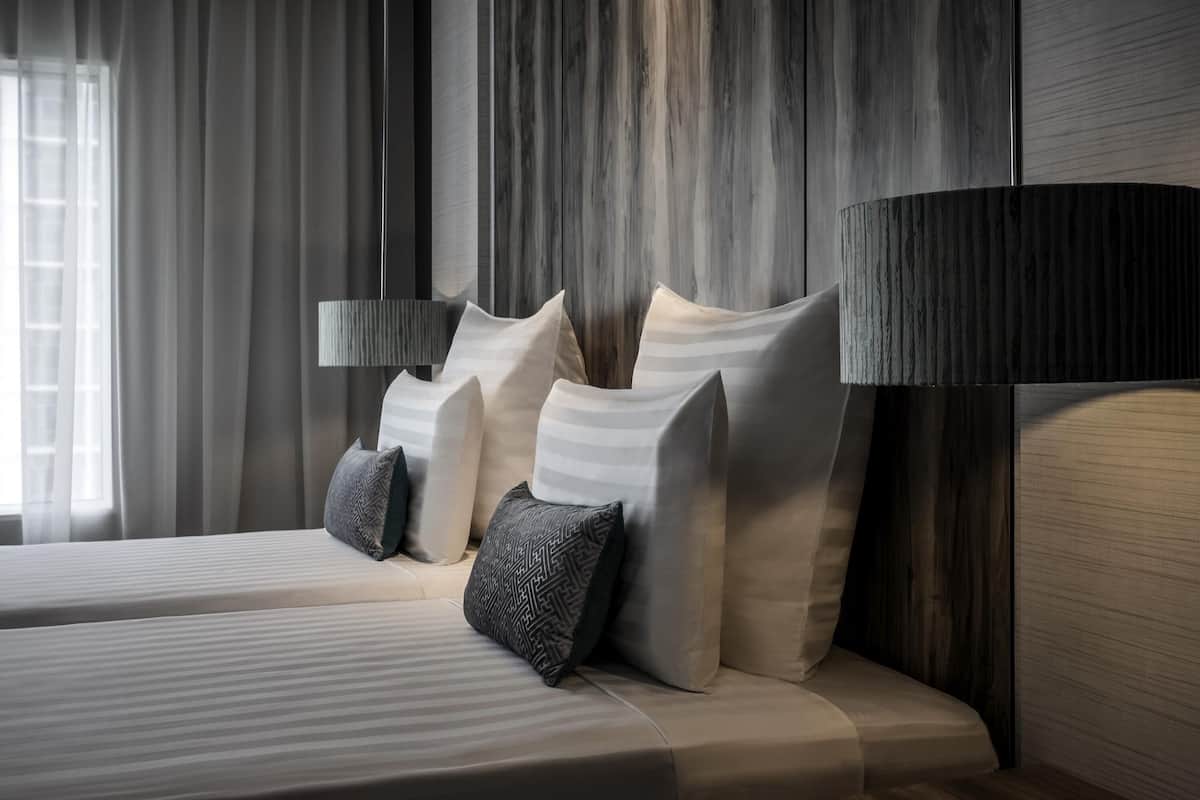 Una habitación de hotel moderna con una cama grande, varios cojines, una lámpara de noche y una ventana con cortinas.
