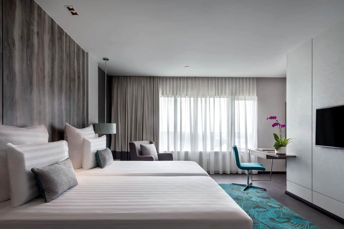 Una habitación de hotel moderna con dos camas, un escritorio, una silla, un televisor y un ventanal con cortinas.