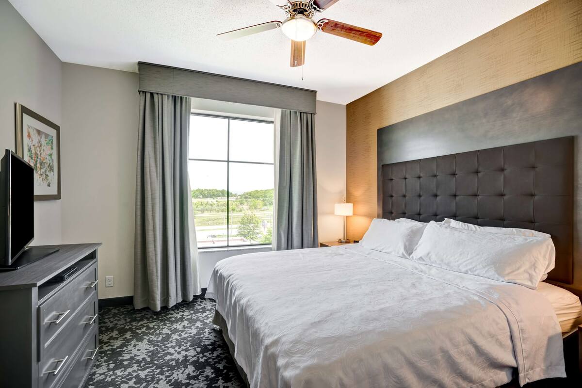 Una habitación de hotel con una cama, un ventilador de techo, un televisor sobre una cómoda, una ventana con cortinas y una lámpara en la mesita de noche.