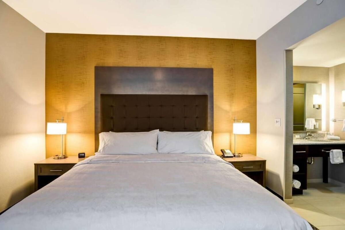 Habitación de hotel con una cama grande, lámparas de noche, mesita de noche, baño con lavamanos y espejo, y un perchero para toallas.