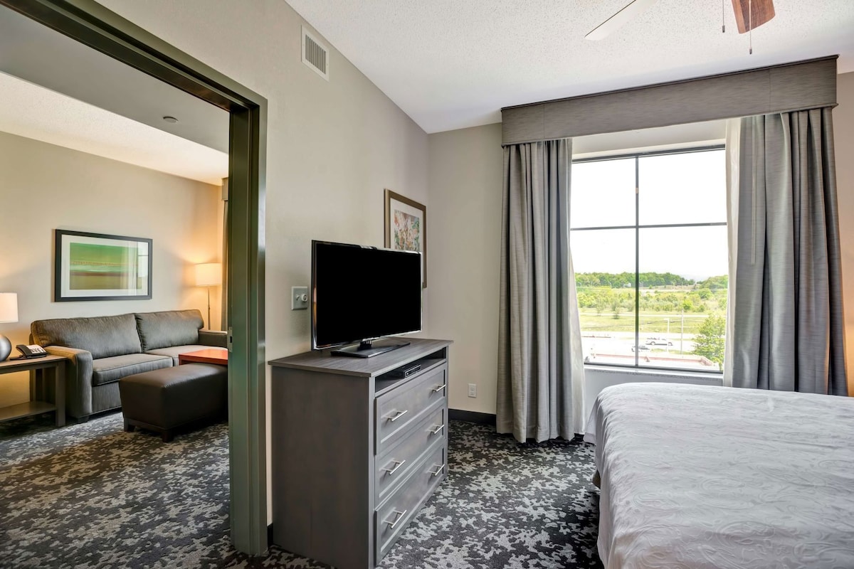 Habitación de hotel con cama, televisor en una cómoda, sofá y un ventanal con cortinas.