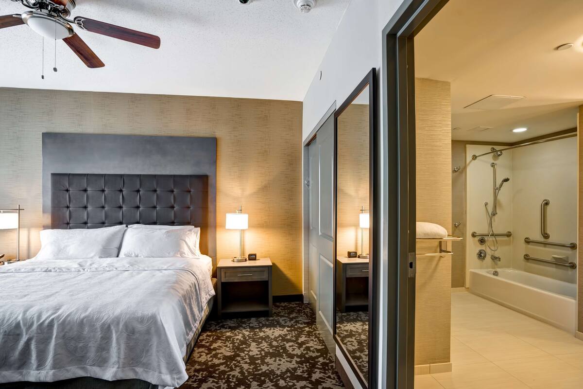 Habitación de hotel con una cama grande, mesitas de noche, ventilador de techo y baño con bañera y ducha.