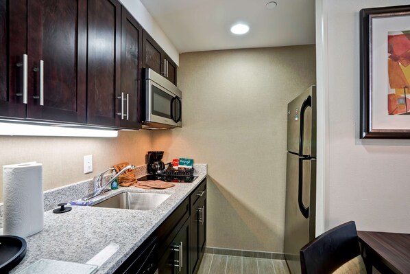 A compact kitchen with dark wood cabinets, a stainless steel microwave, a refrigerator, and a sink.