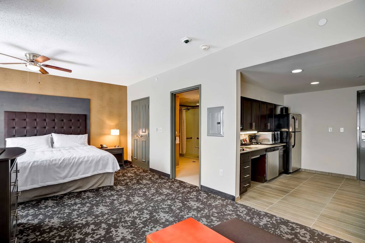 Habitación de hotel con cama, escritorio, silla, ventilador de techo, baño y kitchenette.