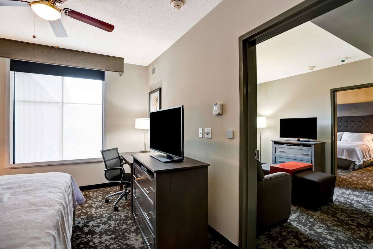 Habitación de hotel con cama, escritorio, silla, televisión y vista a otra habitación.