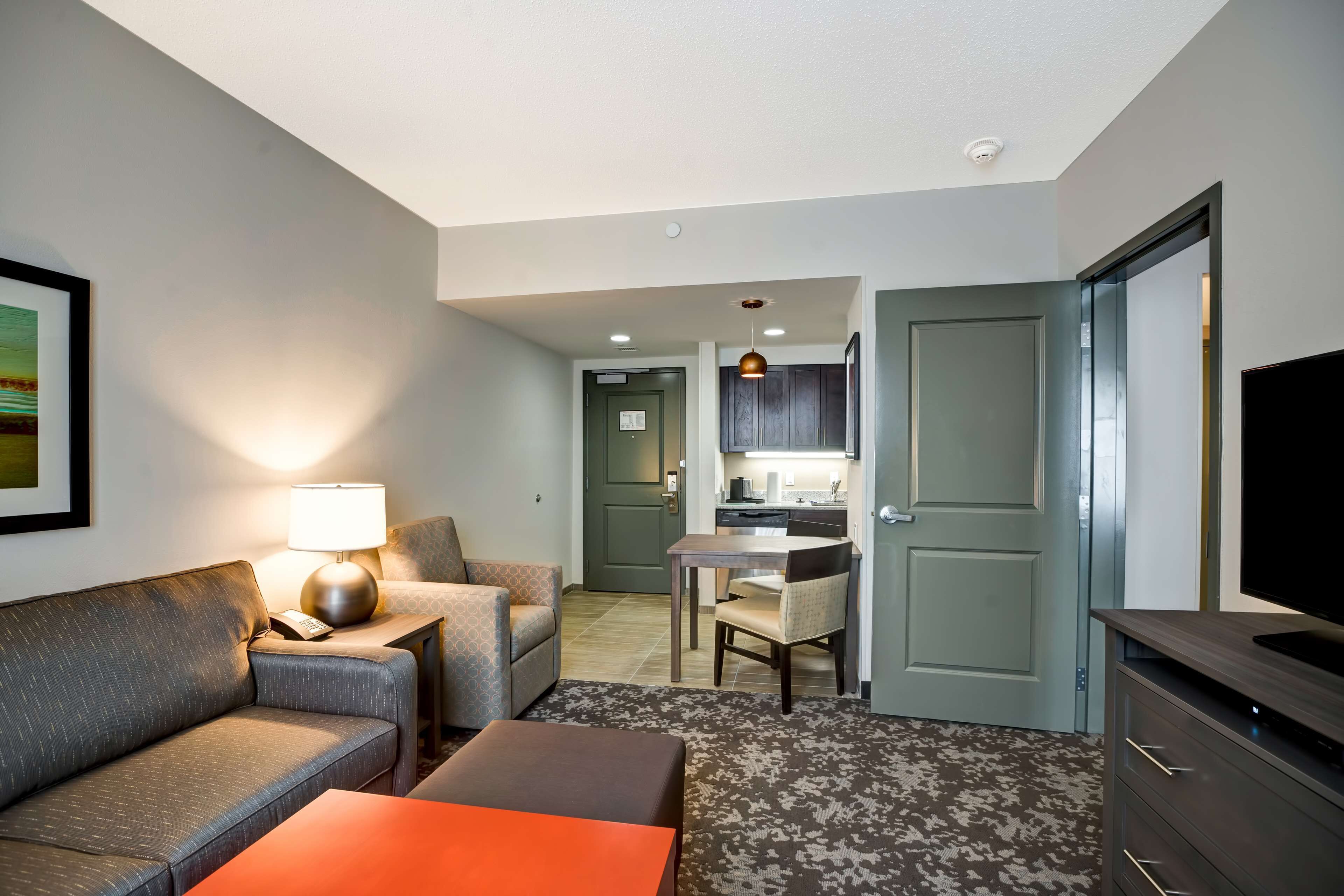 A hotel room with a sofa, a small table, a TV, a lamp, a chair, a kitchen area, and a door.