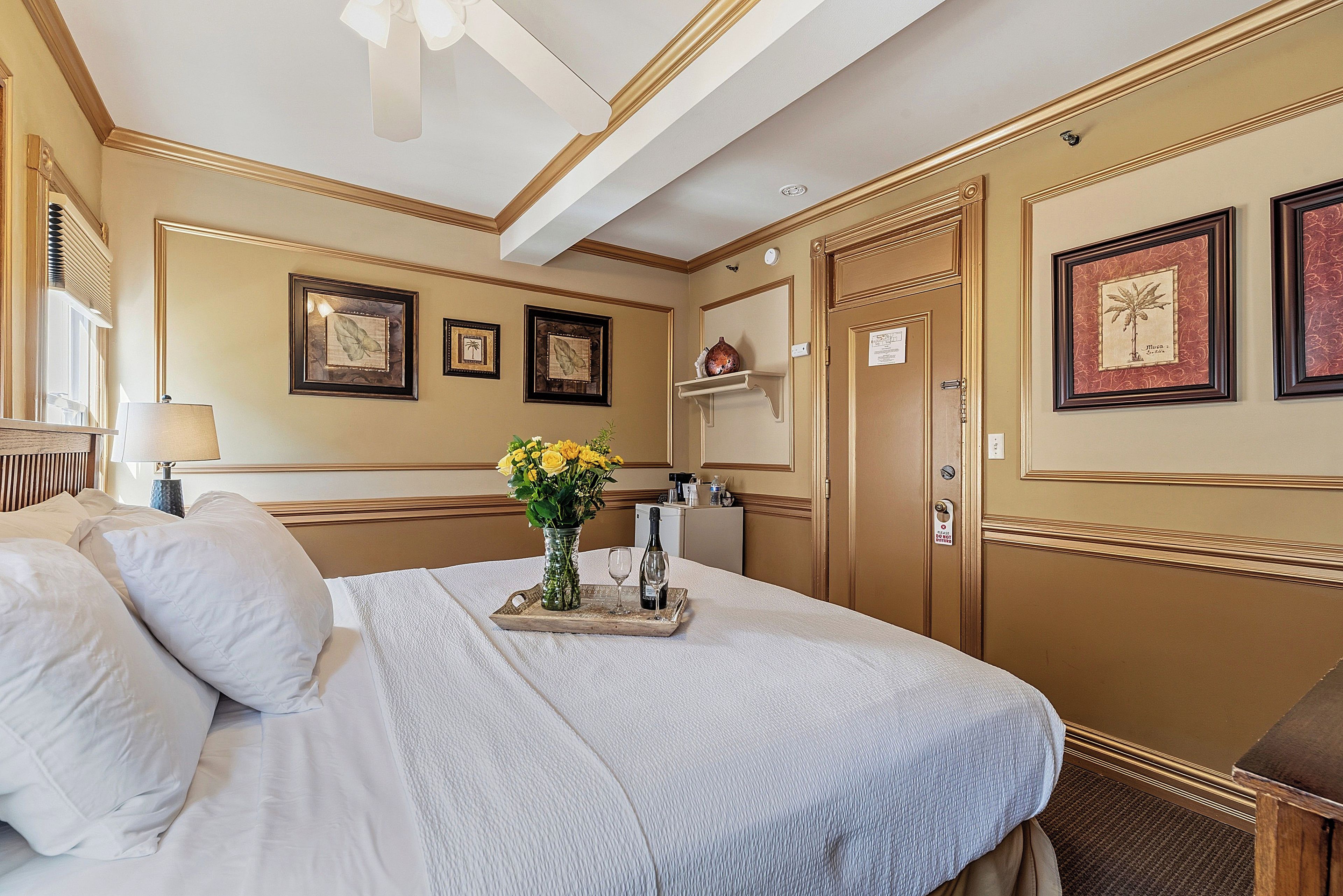 A hotel room with a bed, a vase of flowers, a tray with a bottle and glass, and framed pictures on the wall.