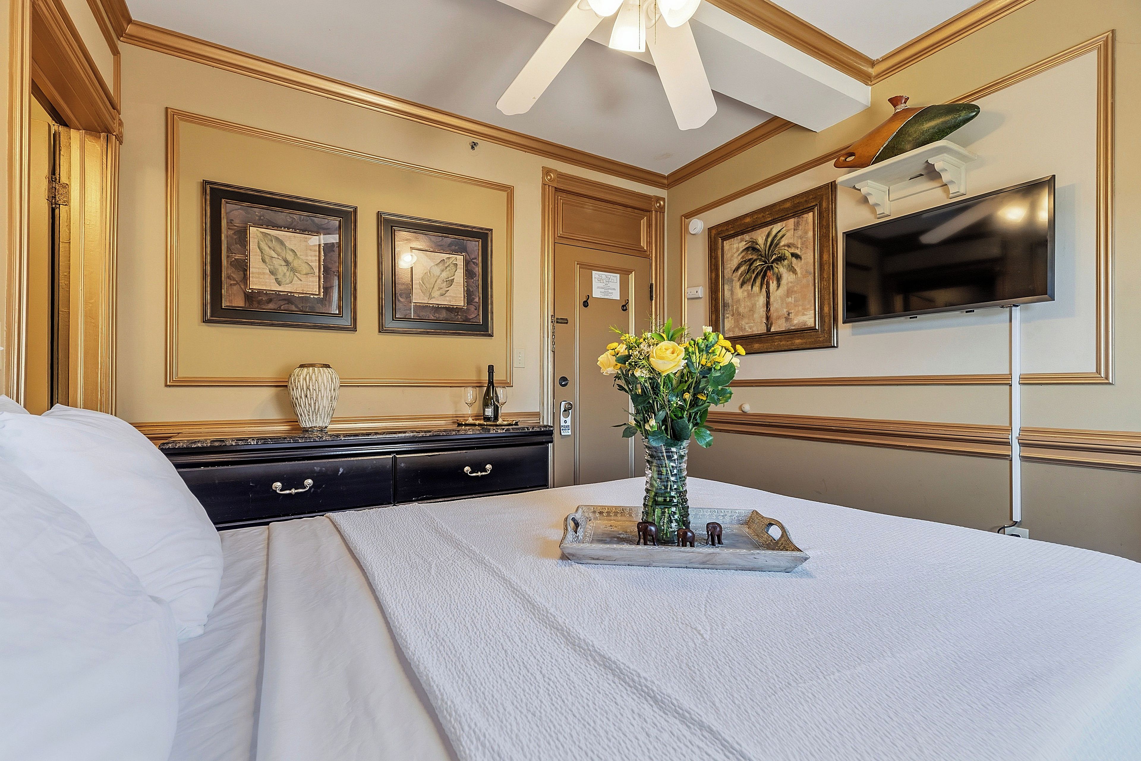 A hotel room with a bed, a dresser, a vase with flowers, a television, and framed artwork on the walls.