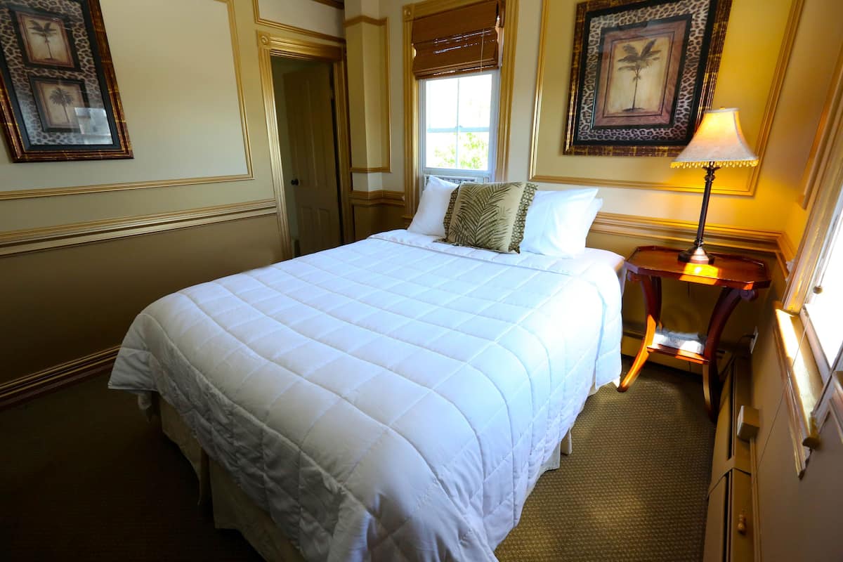 Uma cama bem arrumada com edredom branco, uma mesa de cabeceira com abajur e quadros decorativos nas paredes.