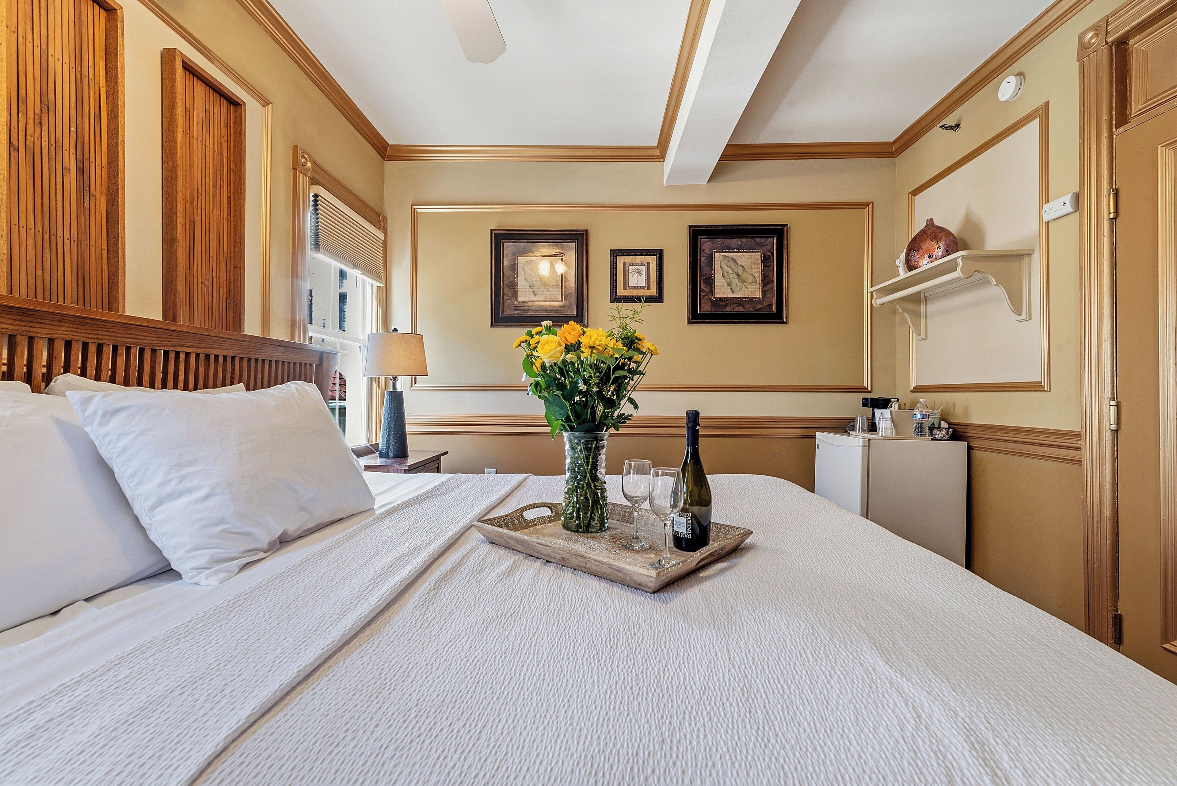 A hotel room with a large bed, a bedside table with a vase of flowers, two wine glasses, and a bottle of wine.