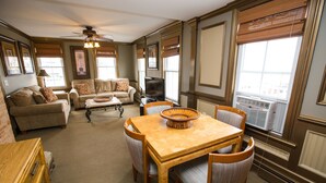 A spacious living room with a wooden dining table, comfortable seating, a television, and large windows with blinds.