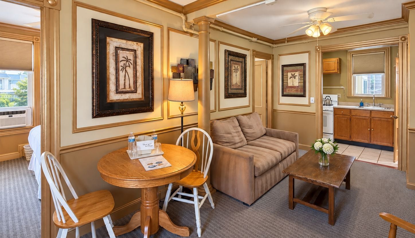 A hotel room with a sofa, a round wooden table, a chair, a small table with a vase, and framed pictures on the wall.