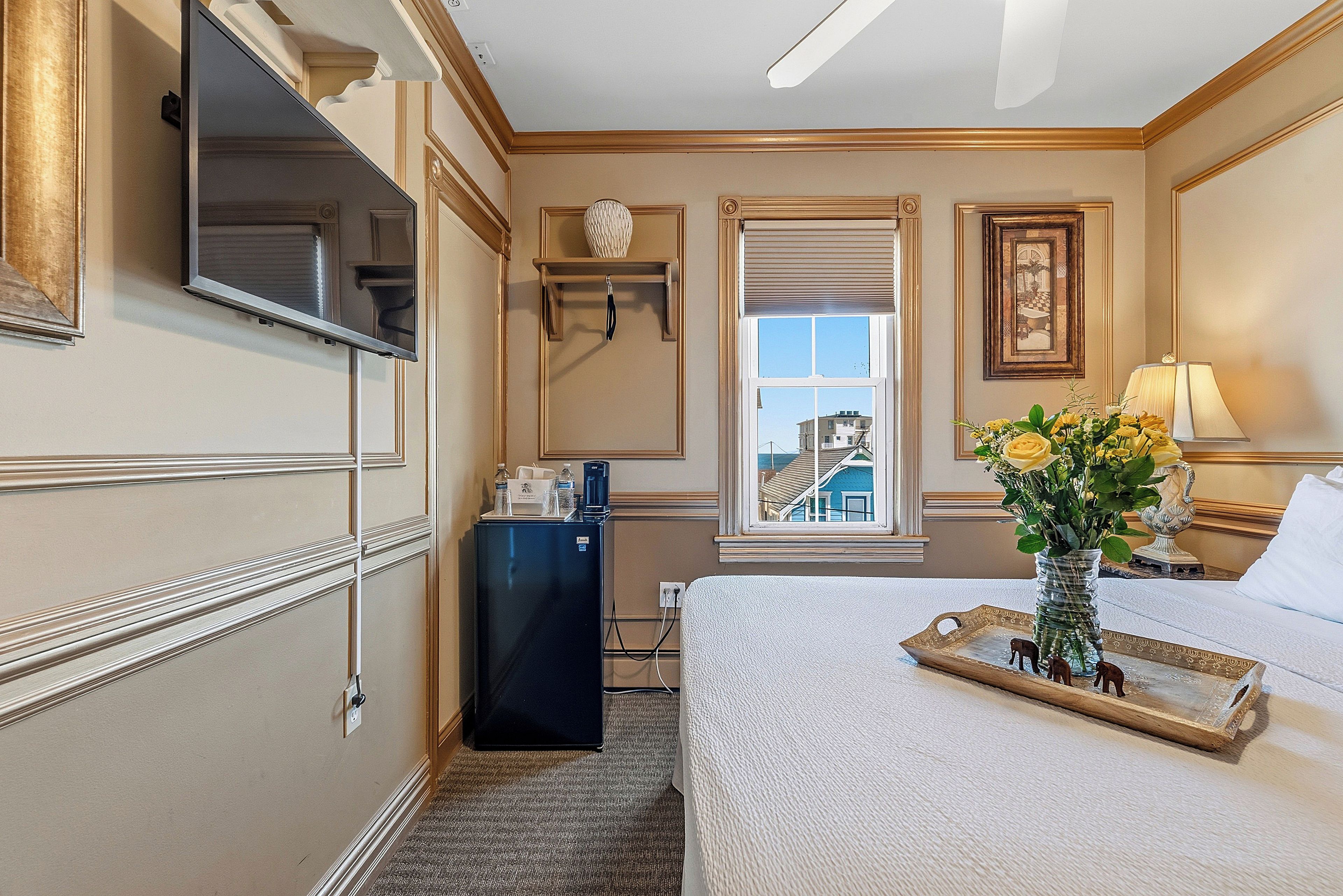 A hotel room with a bed, a desk with a lamp, a window with a view, a small refrigerator, and a decorative vase with flowers.