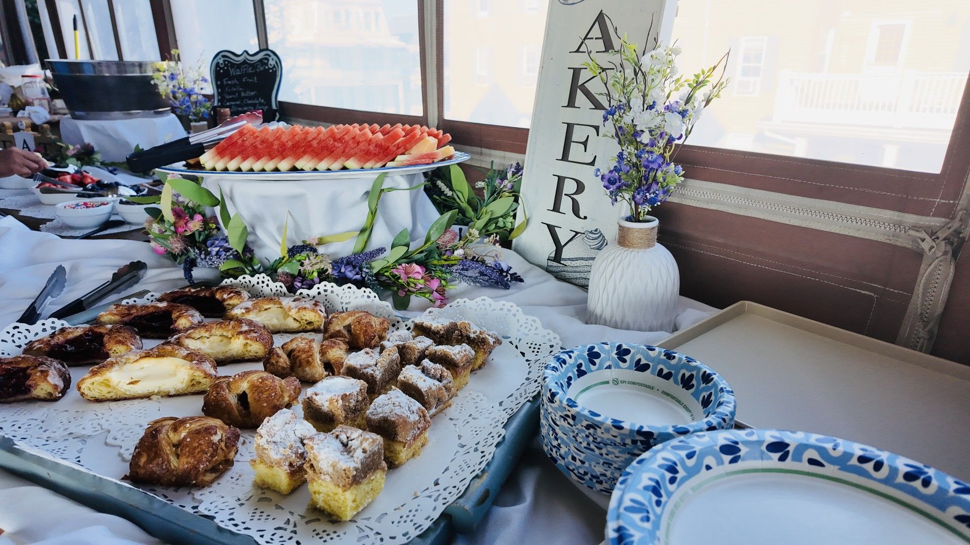 Une table garnie d’une variété de pâtisseries et d’un plat de pastèque, un vase rempli de fleurs et une pancarte sur laquelle est écrit le mot « AKERY ».