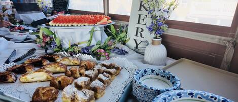 Une table garnie d’une variété de pâtisseries et d’un plat de pastèque, un vase rempli de fleurs et une pancarte sur laquelle est écrit le mot « AKERY ».