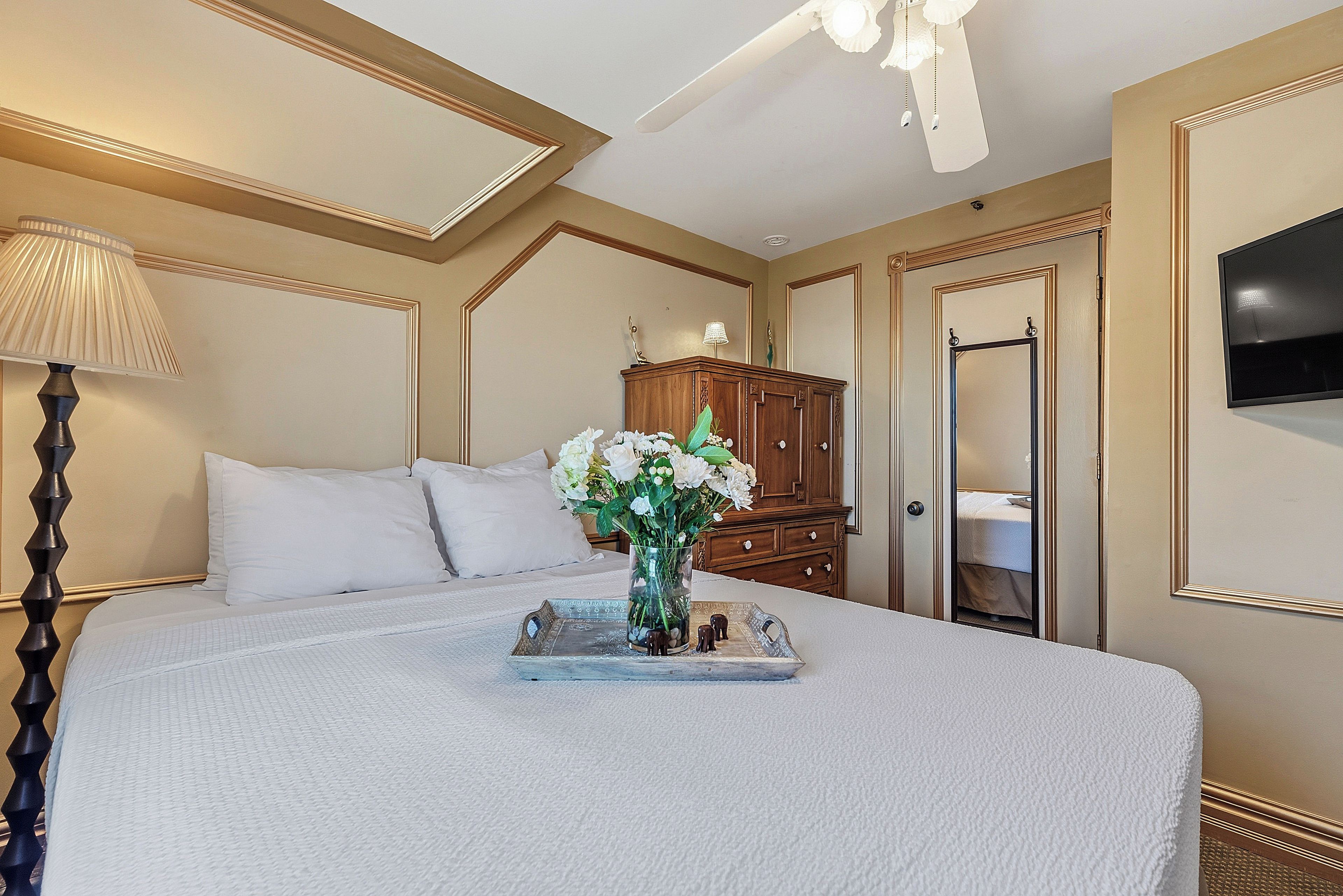 A neatly made bed with a white comforter and pillows, a bedside table with a lamp, a wooden dresser, and a television mounted on the wall.