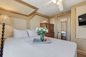 A neatly made bed with a white comforter and pillows, a bedside table with a lamp, a wooden dresser, and a television mounted on the wall.