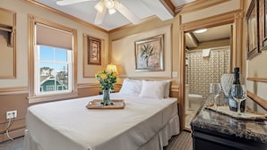 A hotel room with a large bed, a ceiling fan, a window with blinds, a vase with flowers, and a bathroom visible through an open door.