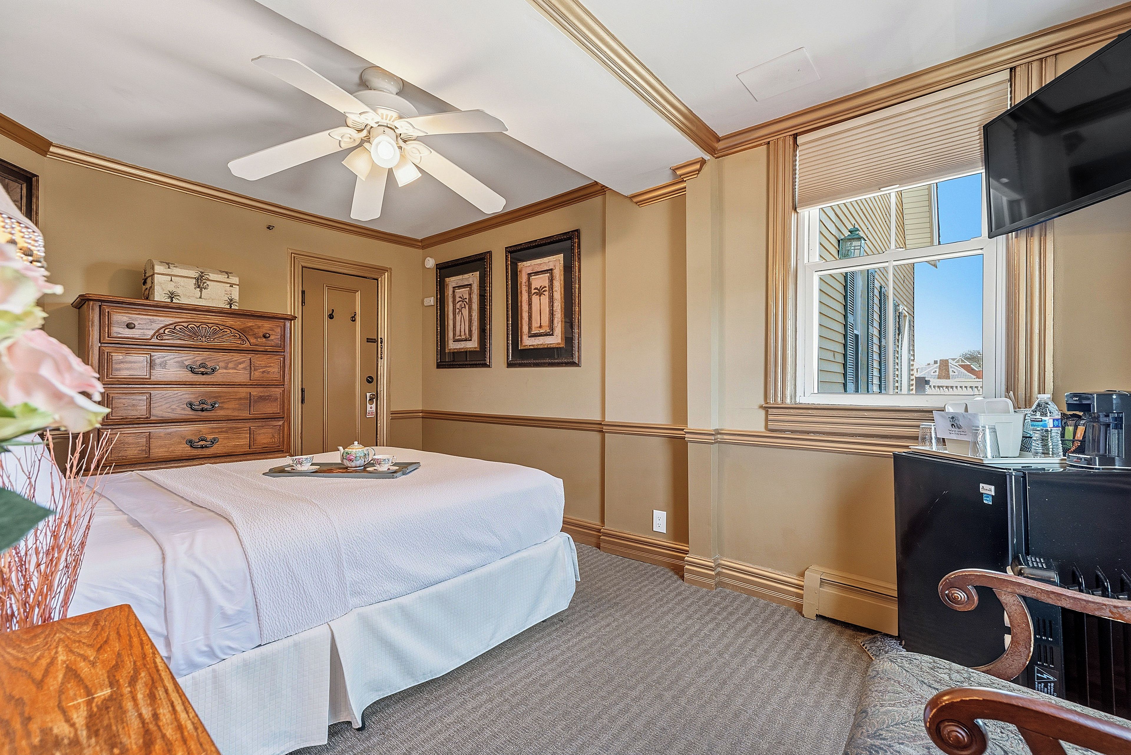 A hotel room with a bed, dresser, ceiling fan, framed pictures, and a window with a view of buildings.