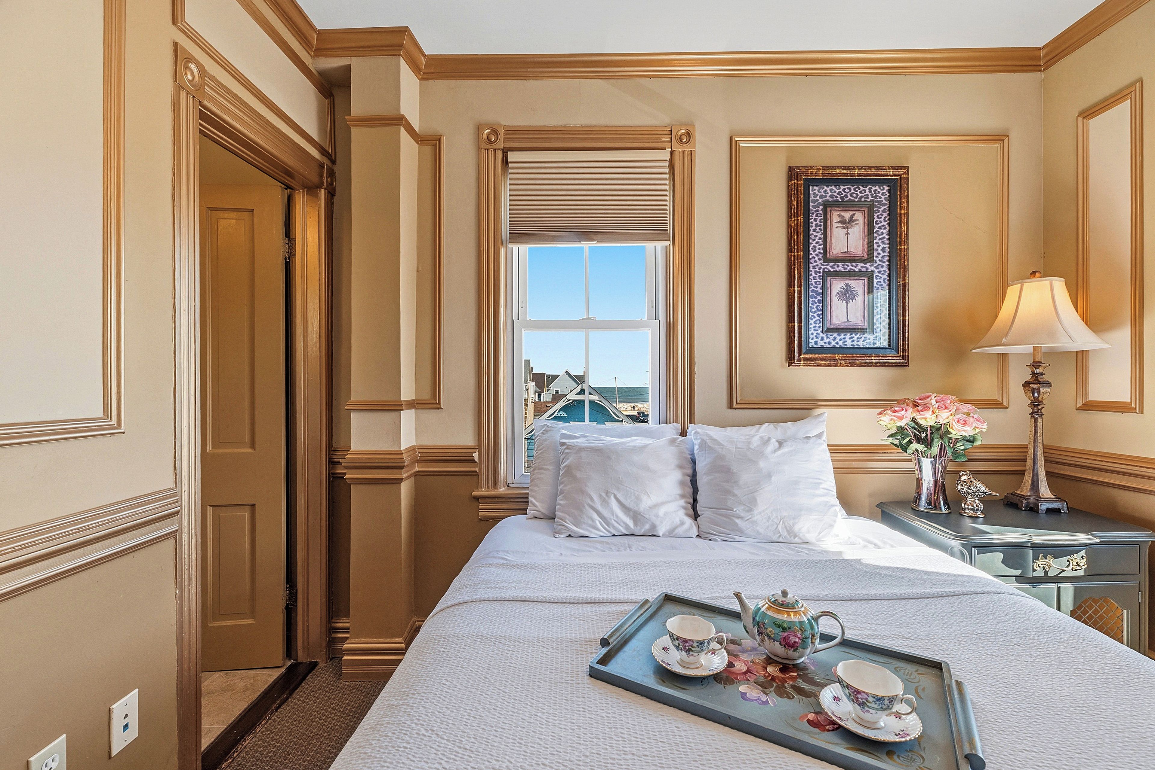 Habitación de hotel con cama, mesita de noche con lámpara, bandeja con juego de té y ventana con vistas a edificios.