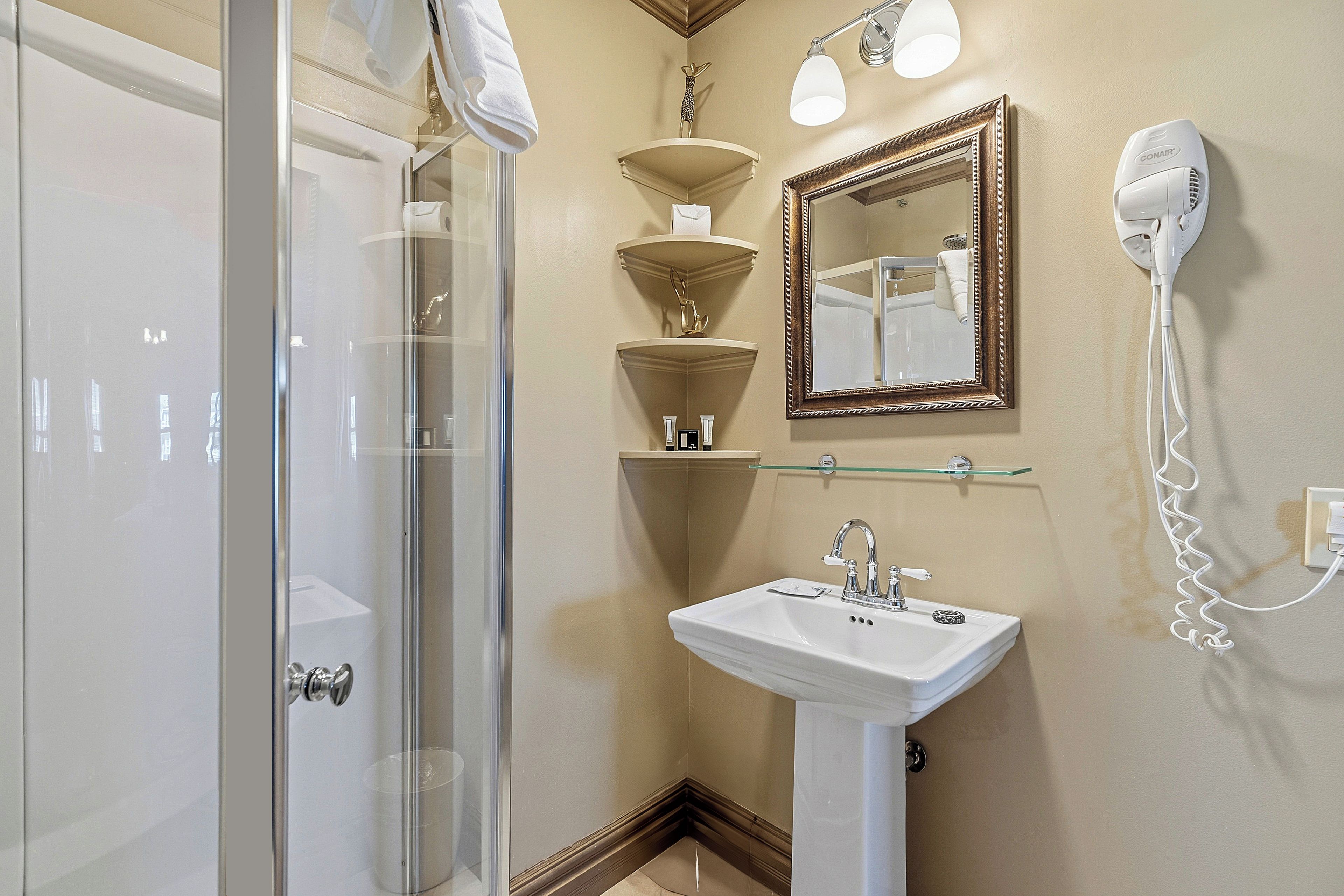 A bathroom with a glass shower enclosure, a white sink, a mirror with a decorative frame, and a hairdryer mounted on the wall.