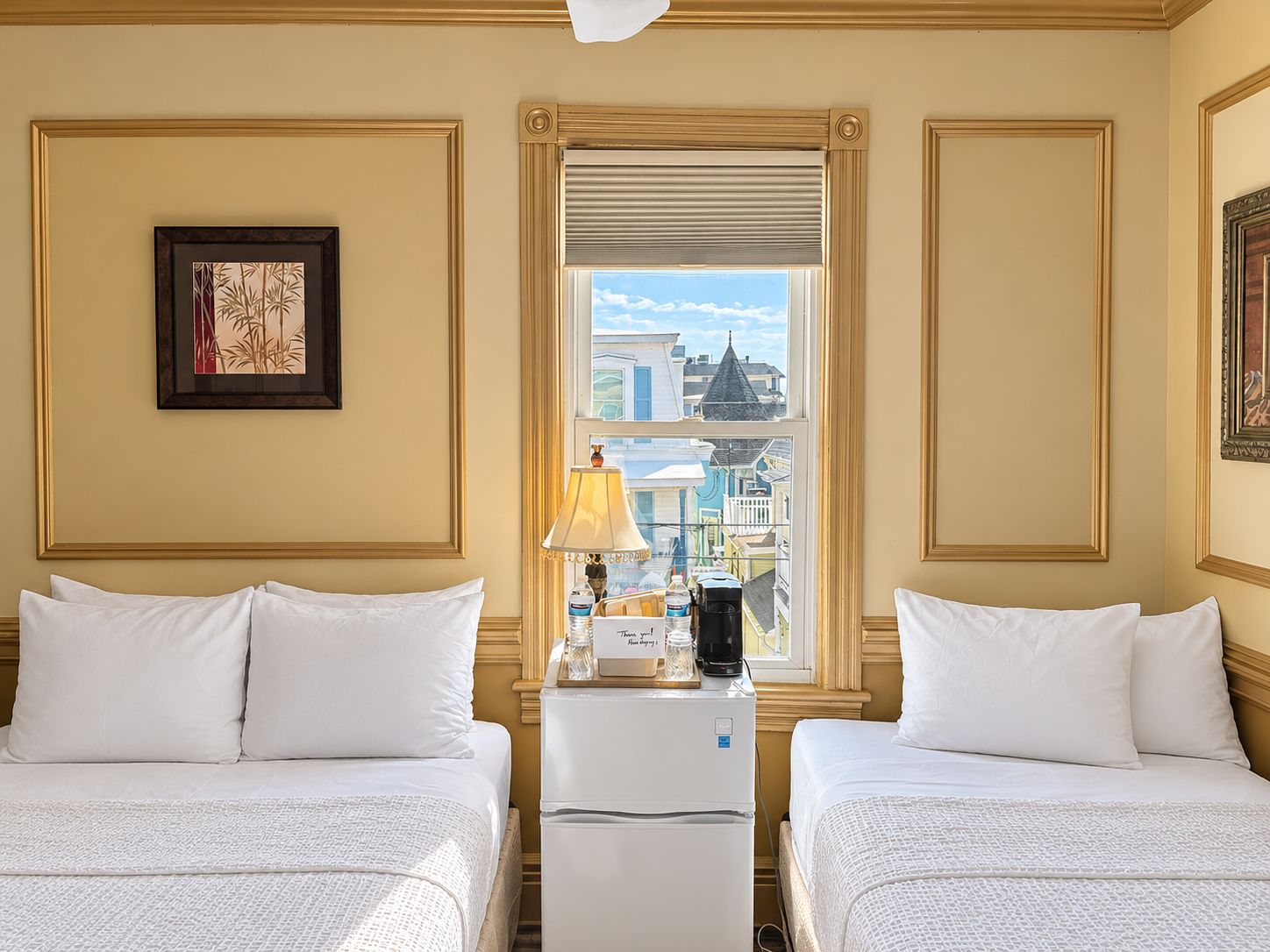 A hotel room with two beds, a window with a city view, a lamp, and a refrigerator.