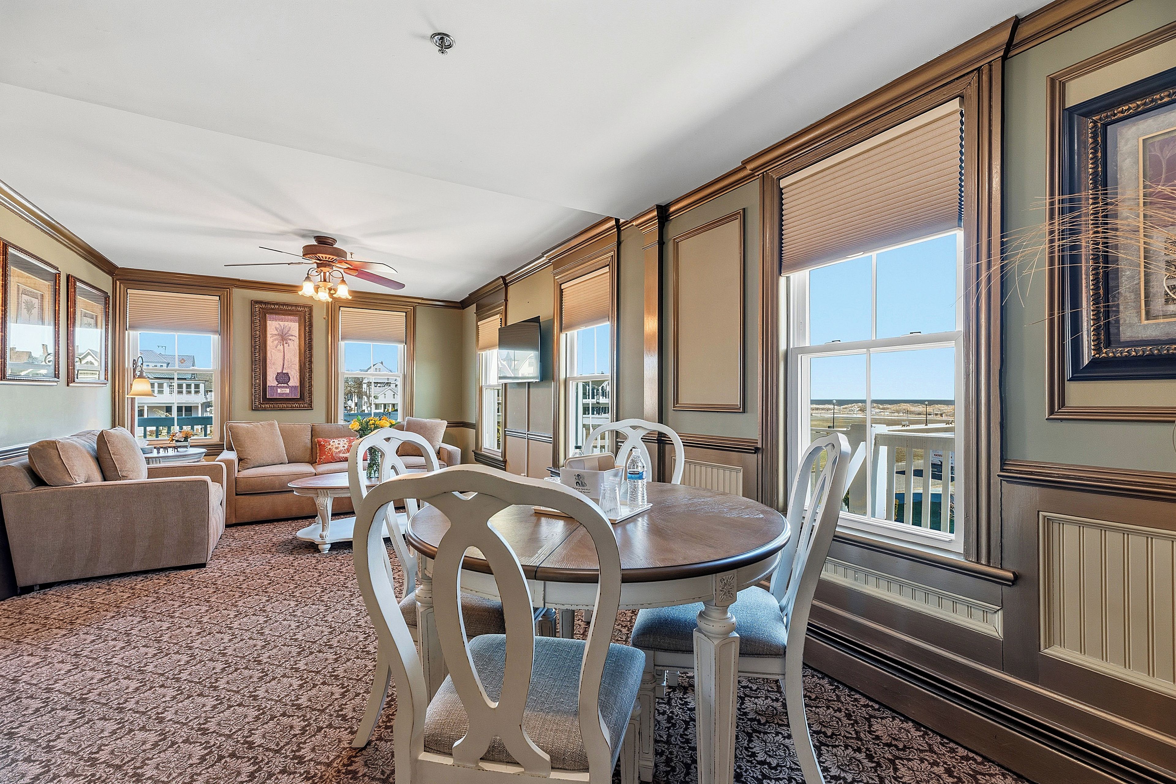 A spacious living room with a ceiling fan, a dining table with chairs, and large windows offering a view of the cityscape.