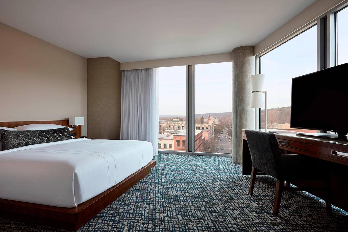 A hotel room with a large bed, a desk with a TV, and a view of the city through large windows.