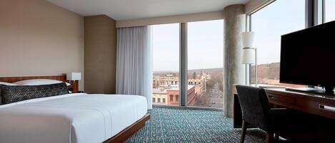 Habitación de hotel con una cama grande, un escritorio con televisor y vistas a la ciudad a través de amplios ventanales.