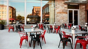 An outdoor cafe with tables and chairs, large windows, and a brick building facade. - Ithaca Marriott Downtown on the Commons (Ithaca)