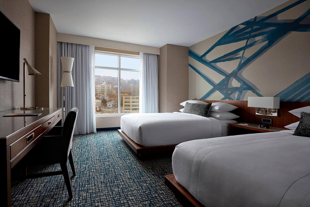A hotel room with two beds, a desk, a chair, a TV, and a large window with curtains.