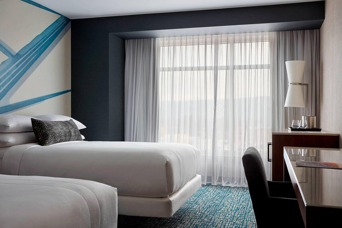 A hotel room with two beds, a desk, a chair, and a large window with sheer curtains.