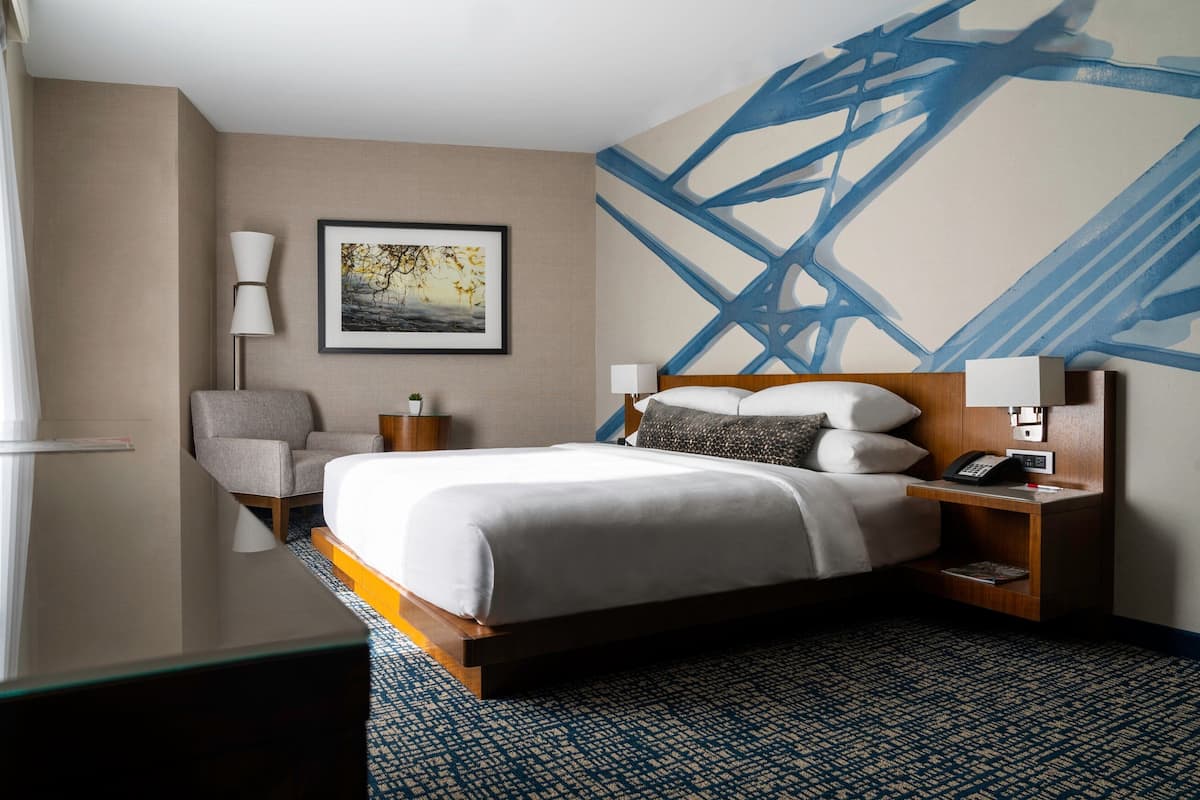 A hotel room with a large bed, a chair, a bedside table with a lamp, and a wall with a blue abstract painting.