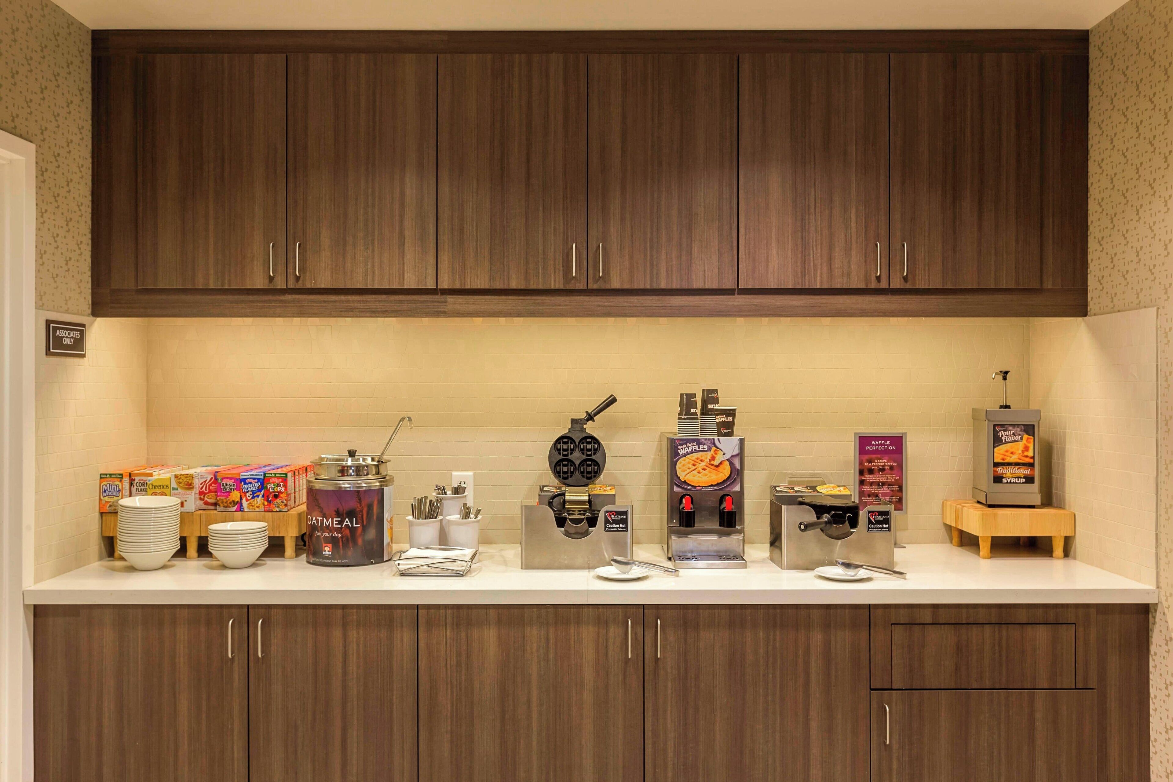 A kitchenette with wooden cabinets, a coffee machine, waffle maker, and a waffle syrup dispenser.