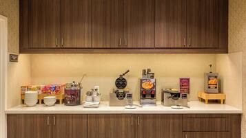 A kitchenette with wooden cabinets, a coffee machine, waffle maker, and a waffle syrup dispenser.