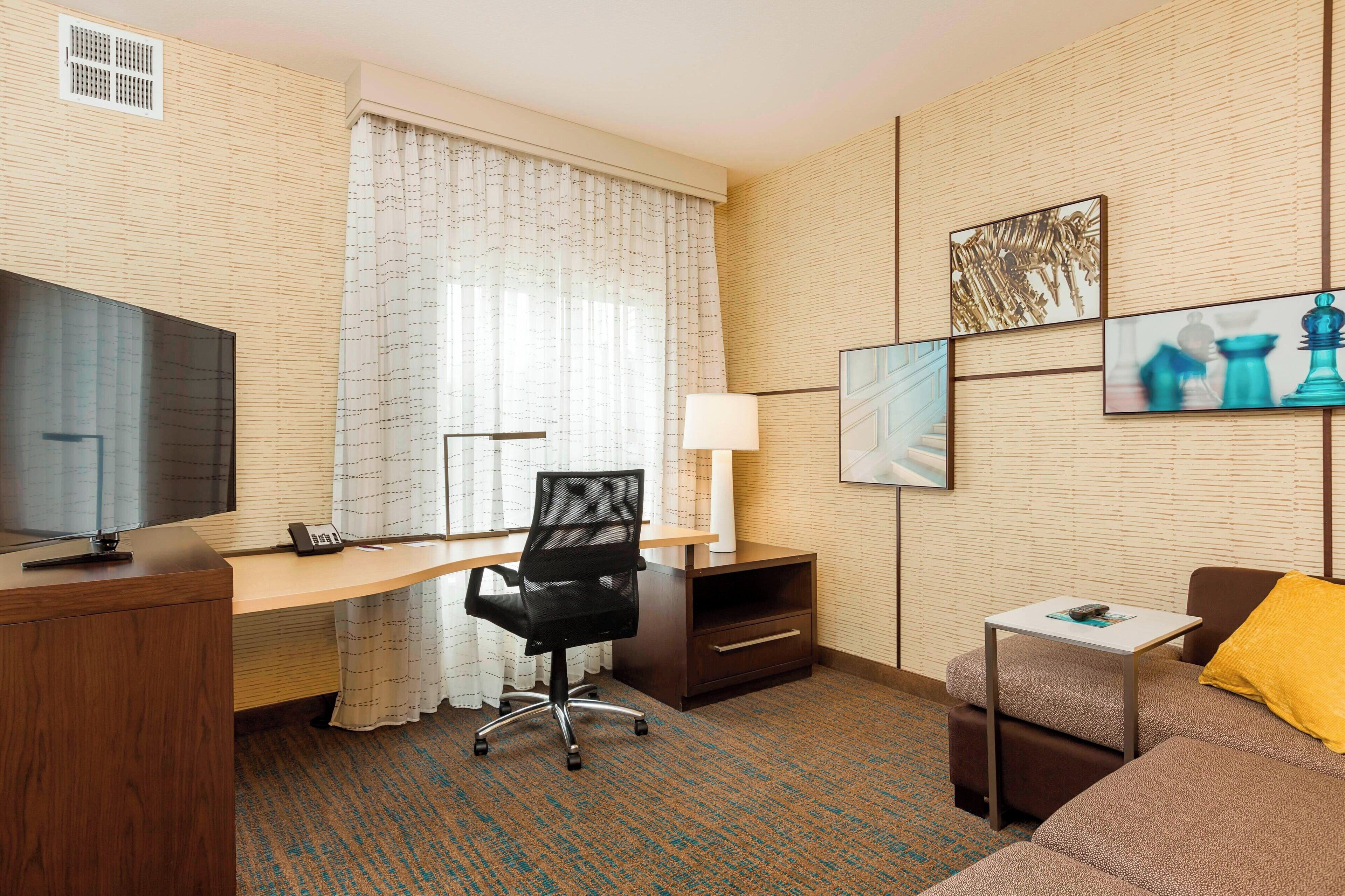 A hotel room with a wooden desk, a black office chair, a television, a lamp, a small table, a sofa, and artwork on the wall.