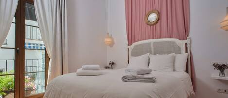 A bedroom with a bed, a window with curtains, a wall-mounted light, and a small shelf with a vase.