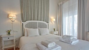 A neatly made bed with white linens and two folded towels, a white headboard, two bedside tables with lamps, and a window with sheer curtains. - Hotel La Morada Mas Hermosa (Marbella)