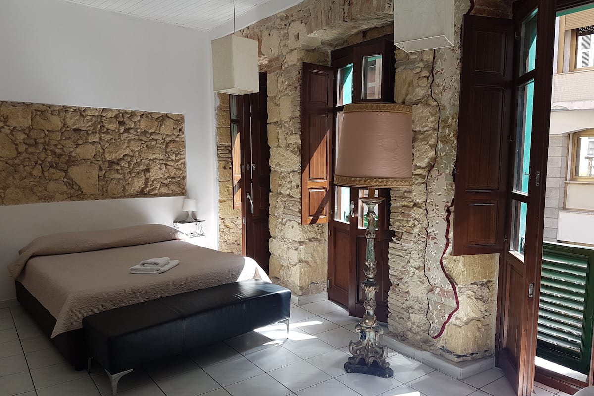 A bedroom with a stone wall, a bed with a headboard, a bedside table with a lamp, and a window with shutters.
