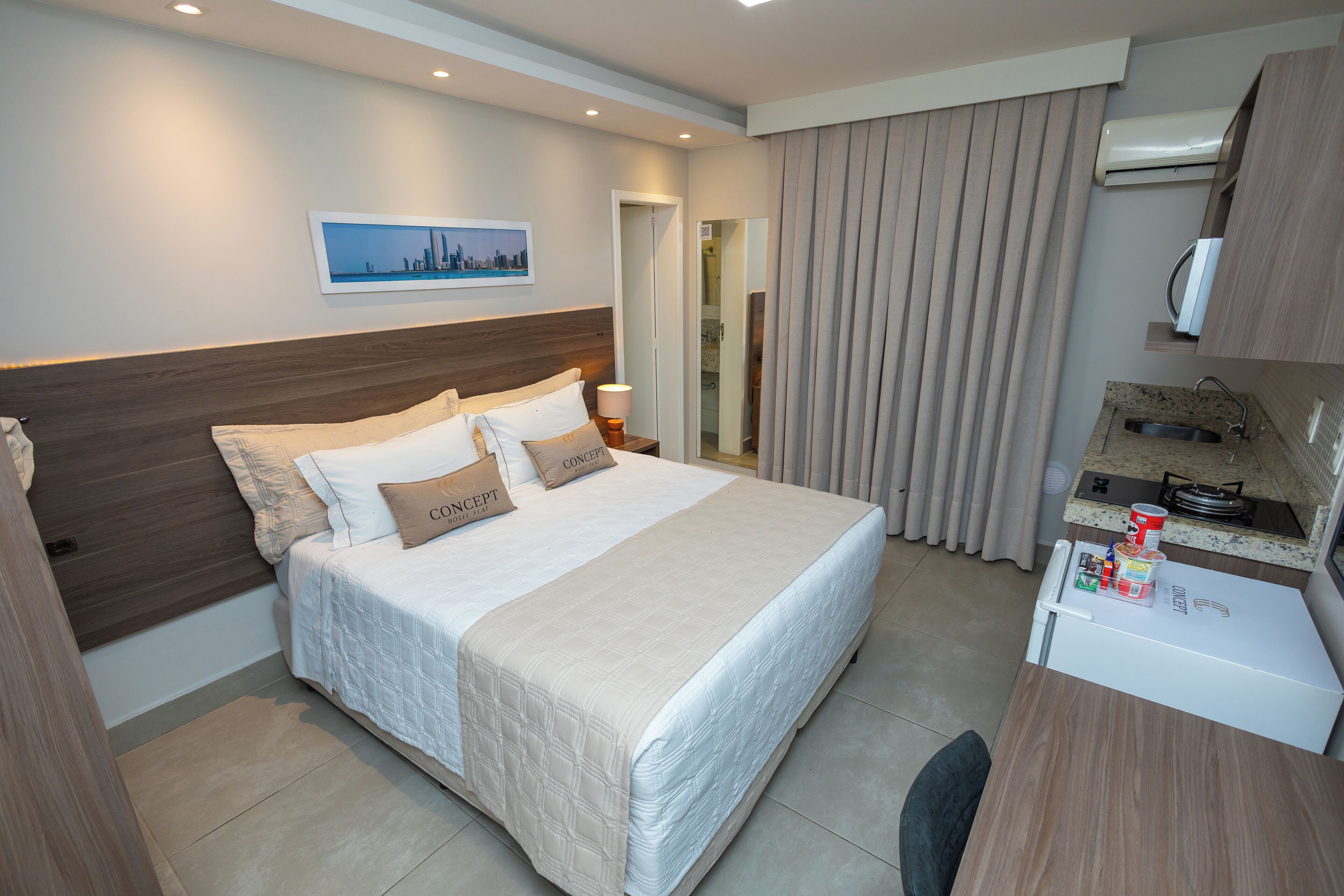 A hotel room with a large bed, a desk with a refrigerator, a window with curtains, and a wall-mounted air conditioner.