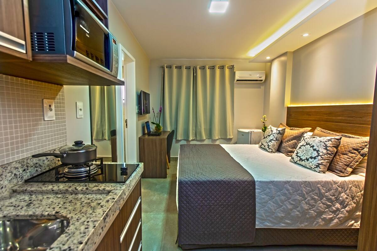 A hotel room with a kitchenette, a bed with a quilted bedspread, a bedside table, and a window with curtains.