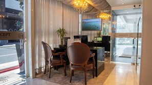 A modern hotel lobby with a reception desk, two wooden chairs, a potted plant, and a television screen displaying a scenic view.