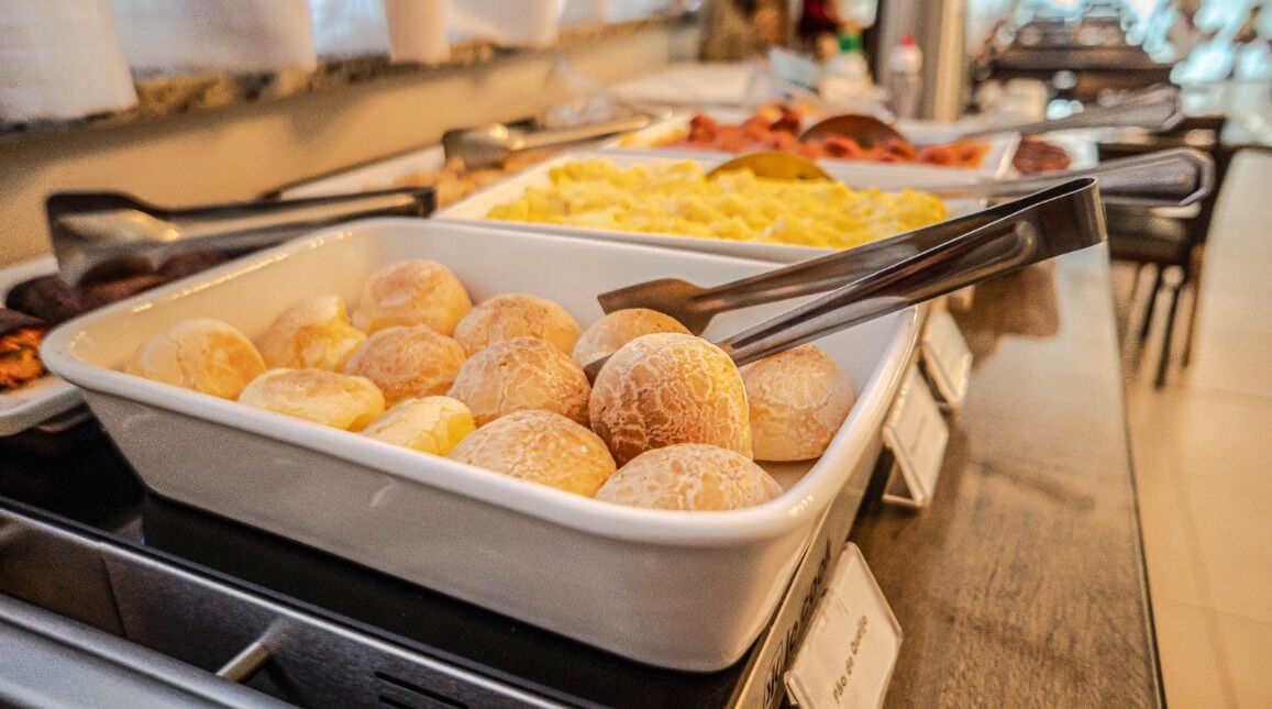 Buffet variado com diversos pratos, incluindo uma bandeja de doces alaranjados e um recipiente com ovos mexidos.