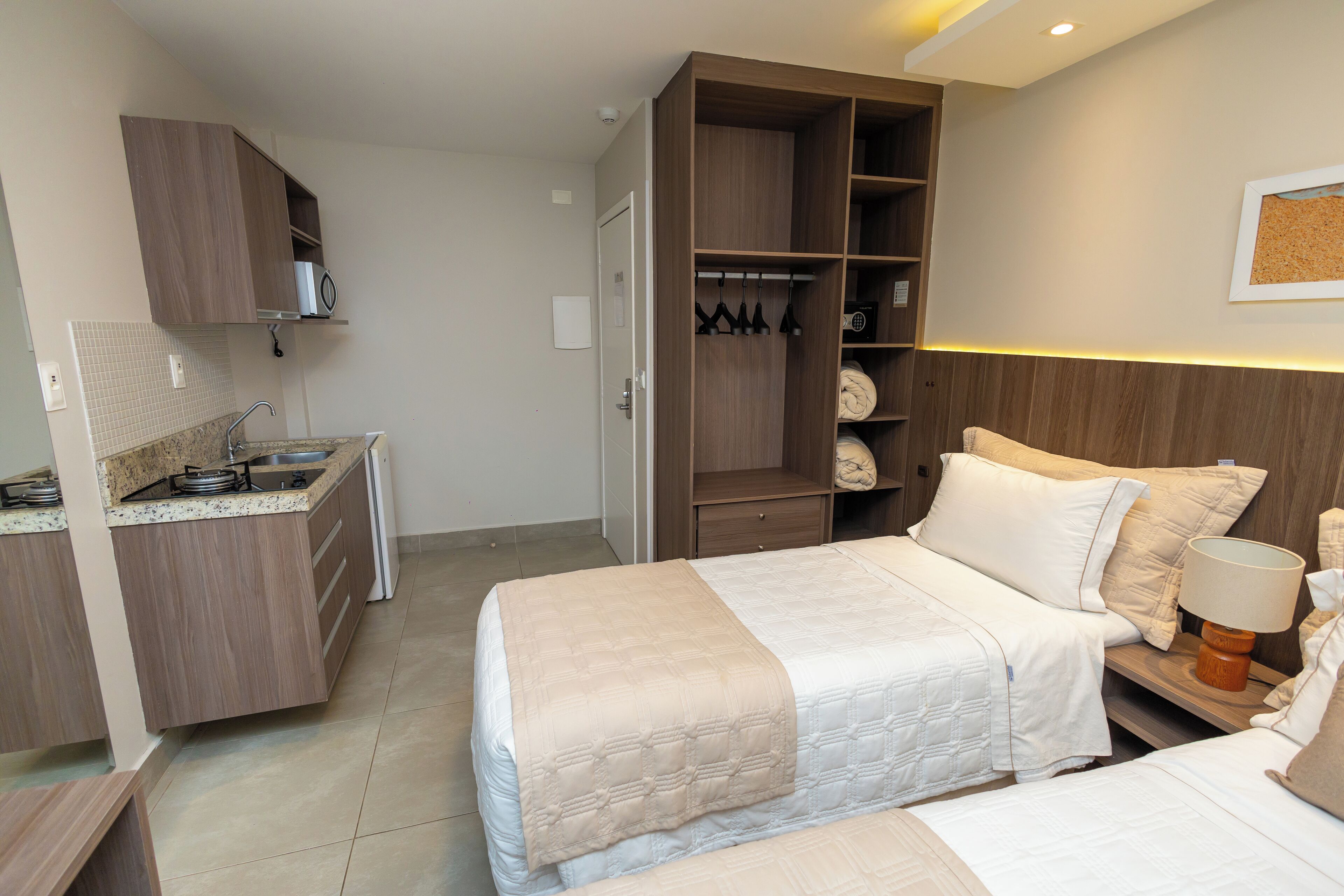 A compact hotel room with a kitchenette, a bed with a white bedspread, a bedside table with a lamp, and a wardrobe.