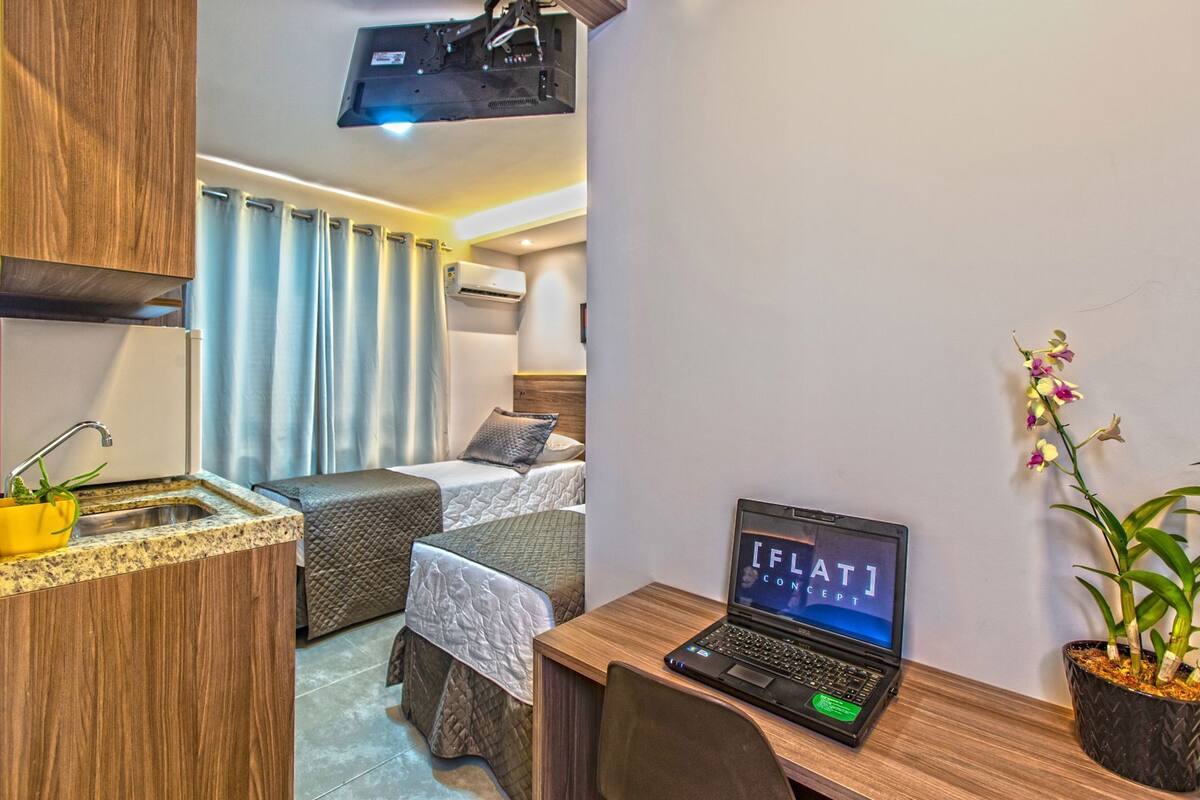 A hotel room with a kitchenette, two beds, a desk with a laptop, and a potted orchid.