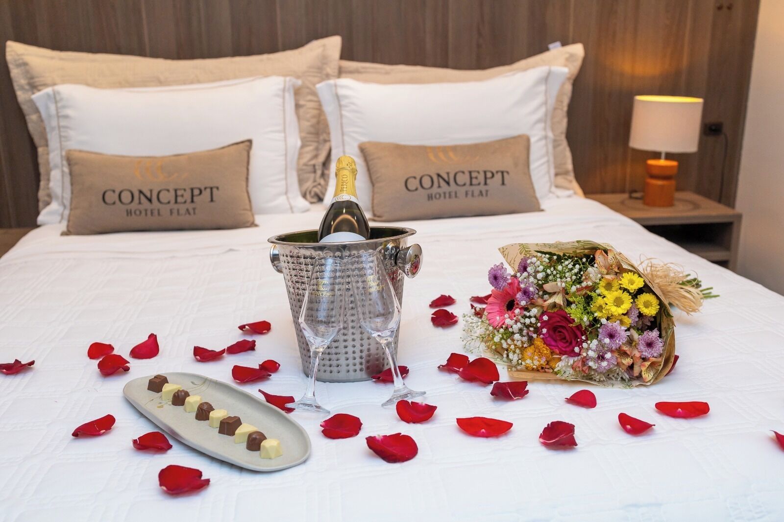 Un lit avec une bouteille de champagne, des verres, des chocolats et un bouquet de fleurs posés sur un couvre-lit blanc.
