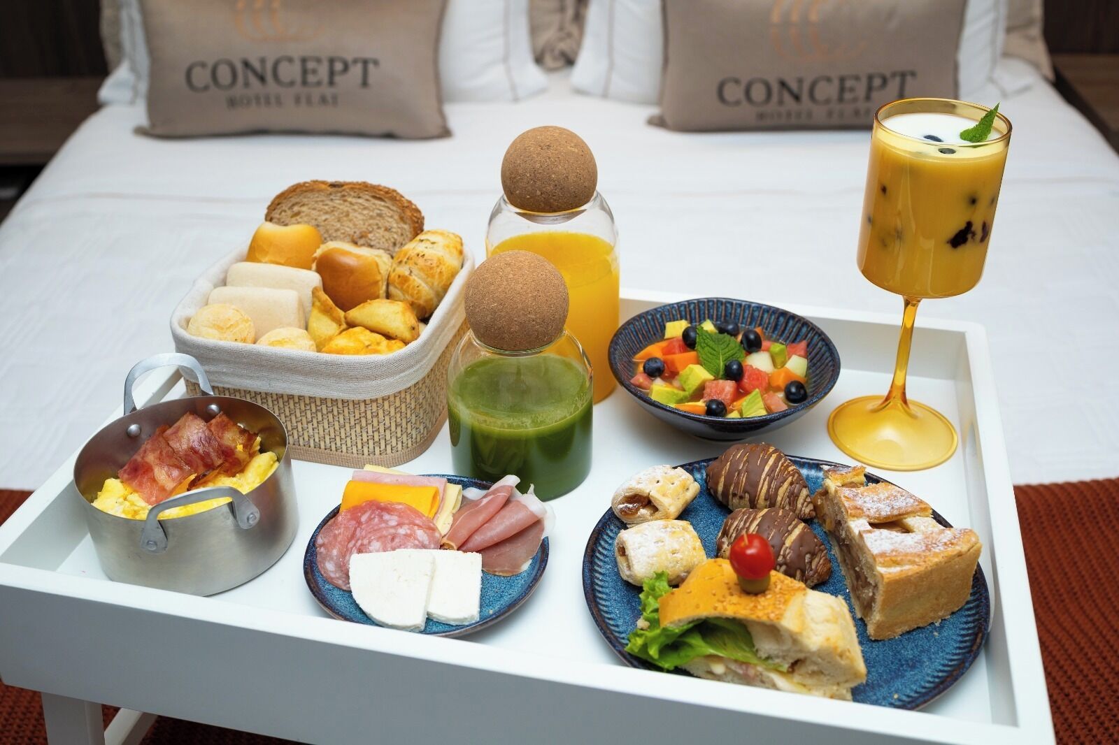 Bandeja de desayuno con diversos alimentos, como huevos, embutidos, bollería y una bebida, sobre una cama con almohadas que dicen 'CONCEPT'.