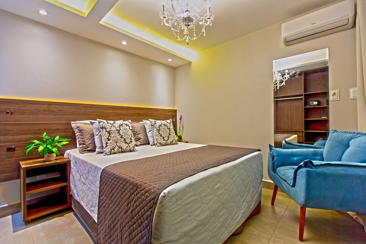A modern bedroom with a large bed, a bedside plant, a blue armchair, and a built-in wardrobe.