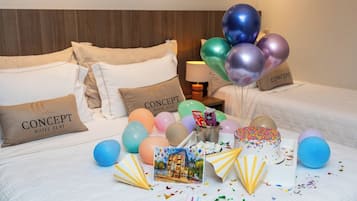 Quarto de hotel com duas camas, cada uma com uma almofada rotulada "CONCEPT HOTEL FLAT". Há balões coloridos, um bolo com granulado e um porta-retrato na cama.