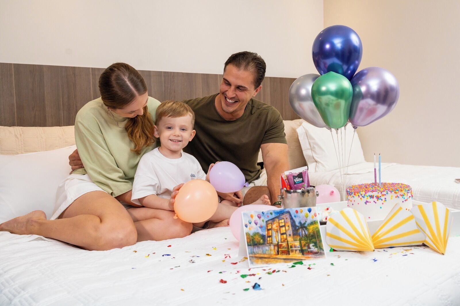 Een gezin op een bed, met ballonnen, een verjaardagstaart en confetti.